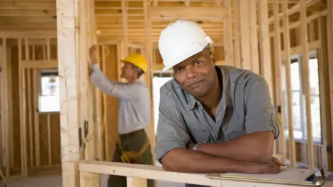 Black male contractor leaning against a building in construction, with a white man in back nailing wood together