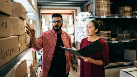Man with glasses pointing at boxes with woman taking notes in a warehouse
